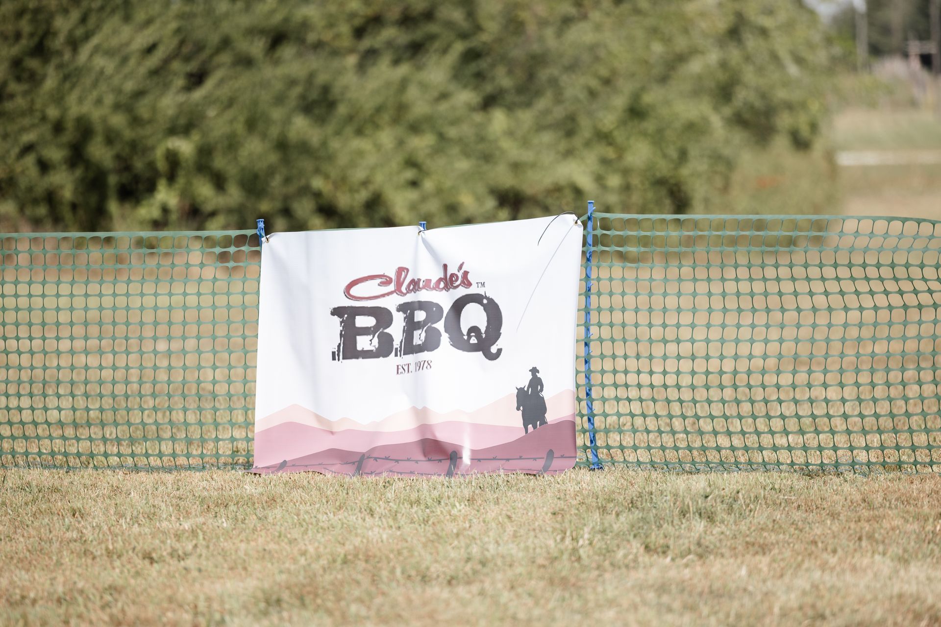Claude's BBQ sign hanging on a green fence in a grassy field with a blurred background of trees.