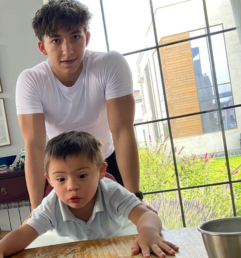 Man in white shirt standing behind child with Down syndrome at a table near a window.