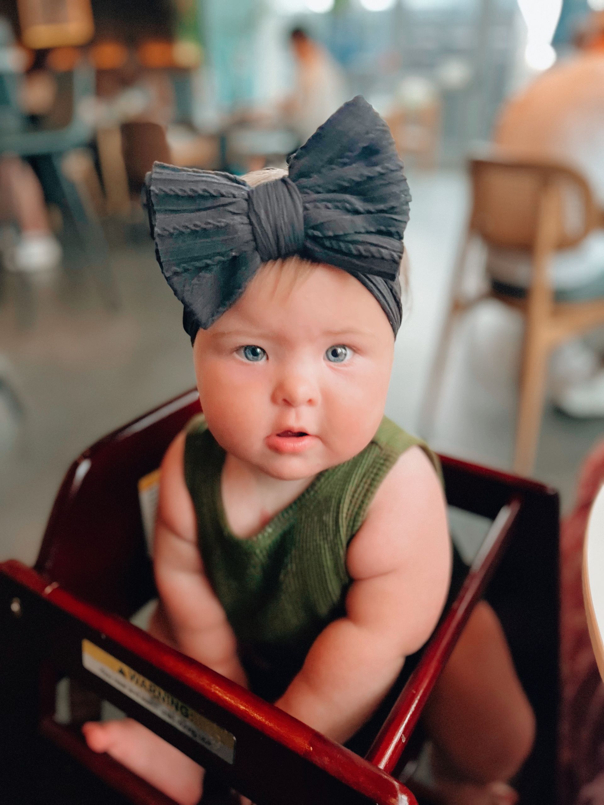Baby with Down syndrome in a high chair wearing a large black bow headband, olive green tank top, looking forward.