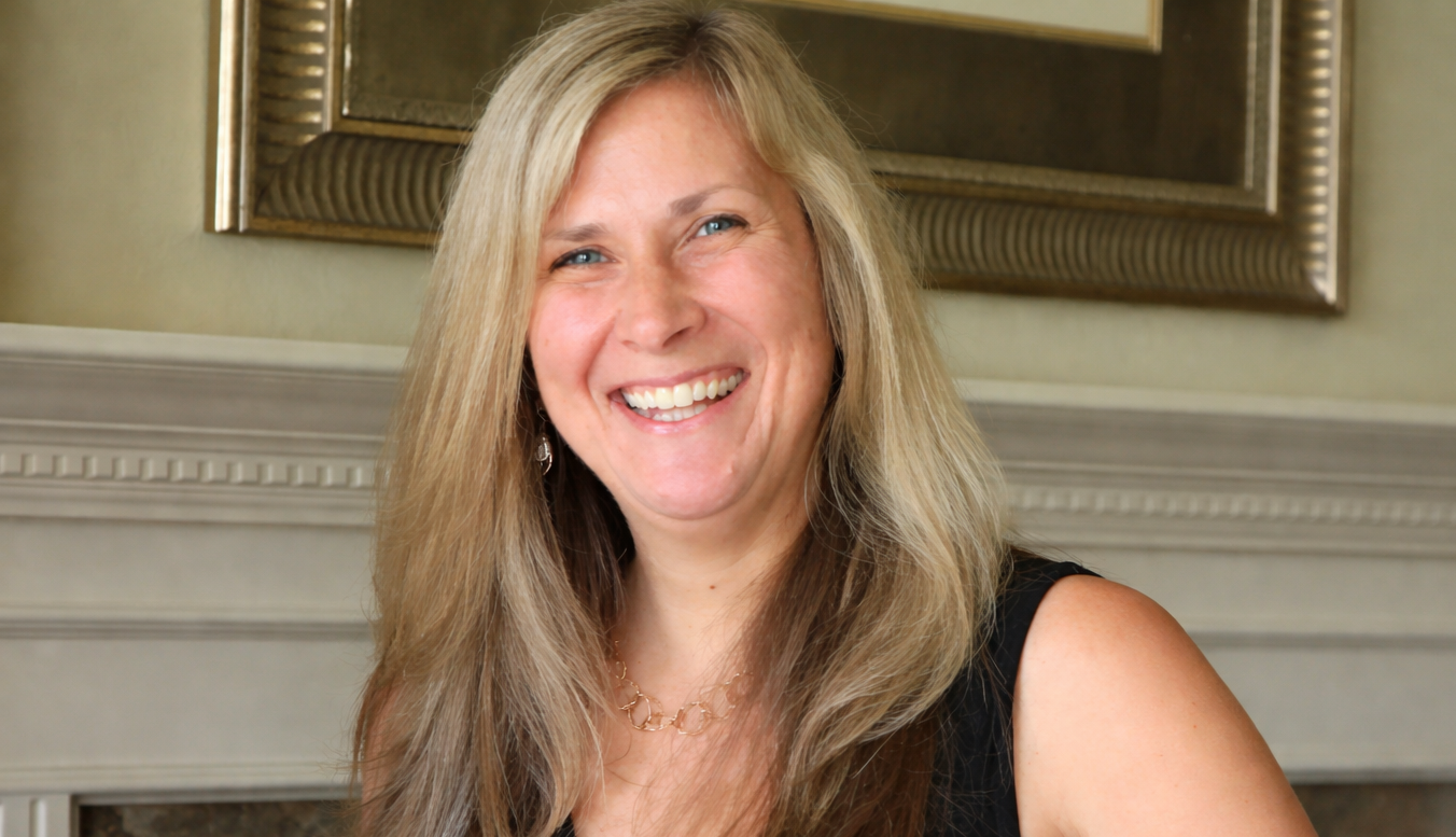 Woman with long dark hair, wearing a gray zip-up sweatshirt, smiles at the camera against a plain background.