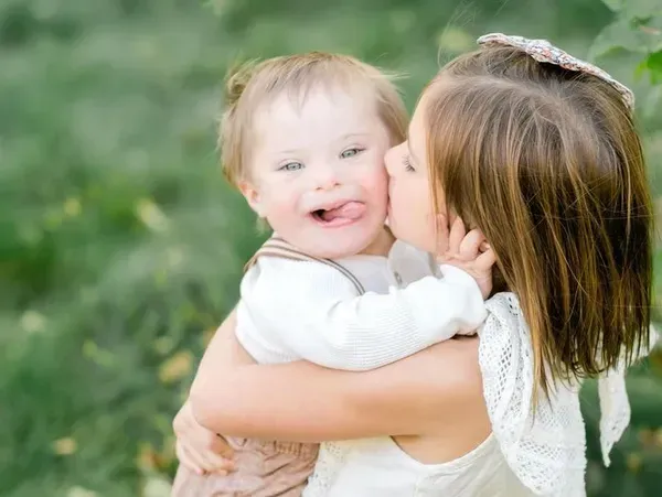 Young girl hugging and kissing a smiling child with Down syndrome outdoors.