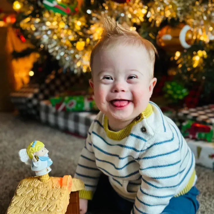 Child with Down syndrome smiles, sticking out tongue, in front of Christmas tree.