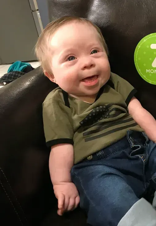 Smiling baby "iron will" with Down syndrome wearing a green shirt and jeans, sitting on a brown couch.