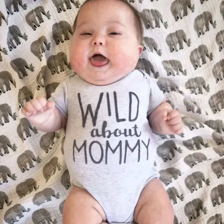 Baby with Down syndrome in gray onesie with "Wild About Mommy" text, lying on elephant-print blanket, mouth open.