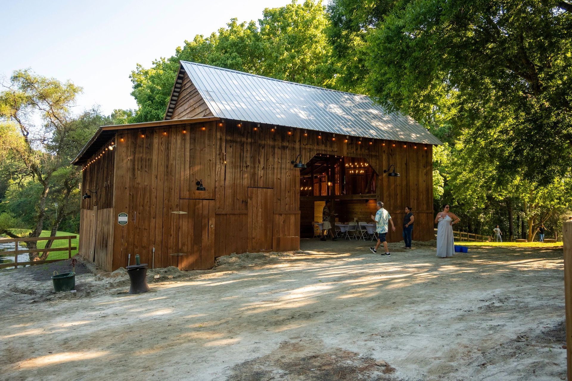 Rustic wooden barn with a metal roof in a sunny clearing, with a few people standing nearby