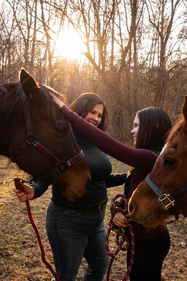 Two women pet two horses in a wooded field at sunset