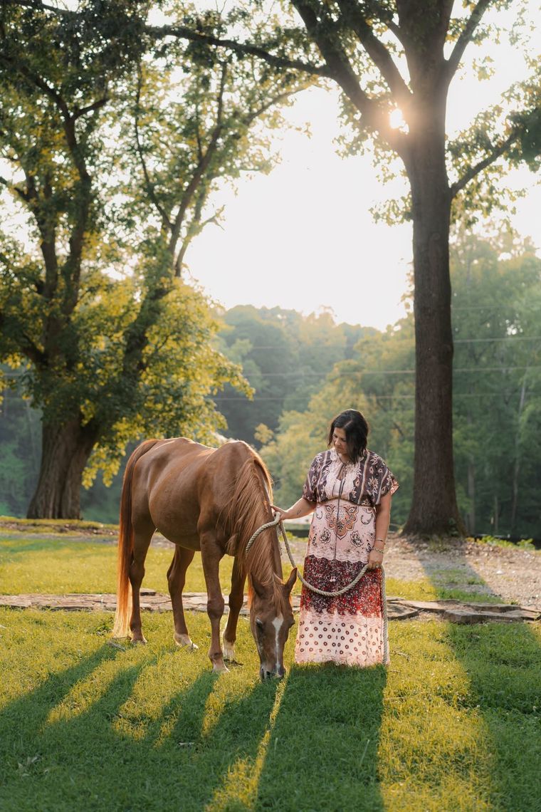 Person standing beside a grazing horse in a sunlit park with tall trees and long shadows