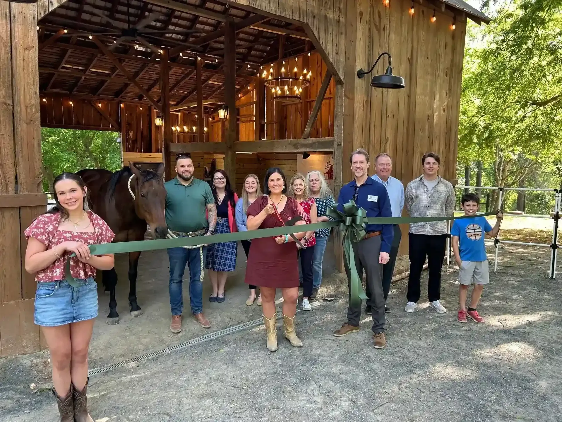 Group standing in front of a rustic barn, holding a long ribbon for a ceremonial opening.