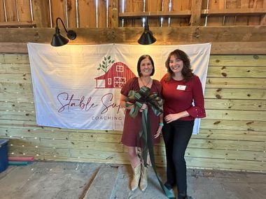 Two women posing indoors before a white banner, holding a bouquet and smiling