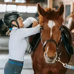 Person adjusting a brown horse’s saddle in an indoor riding arena