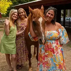 Three people standing with a brown horse under a wooden shelter, smiling in a garden setting.