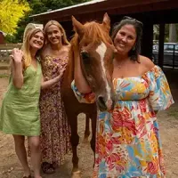 Three people standing with a brown horse under a wooden shelter, smiling in a garden setting.
