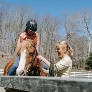 Two people smiling with a horse at an outdoor riding ring, leafless trees in the background