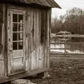 Weathered wooden cabin beside a fenced, muddy field under an overcast sky