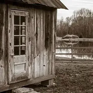 Weathered wooden cabin beside a fenced, muddy field under an overcast sky