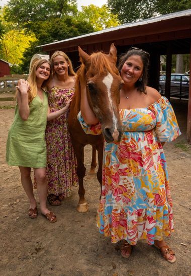 Three women standing with a brown horse in a barnyard, smiling in colorful dresses.