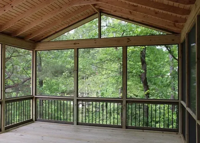 A screened-in porch with wood paneling and railings looking out onto a dense, green forest.