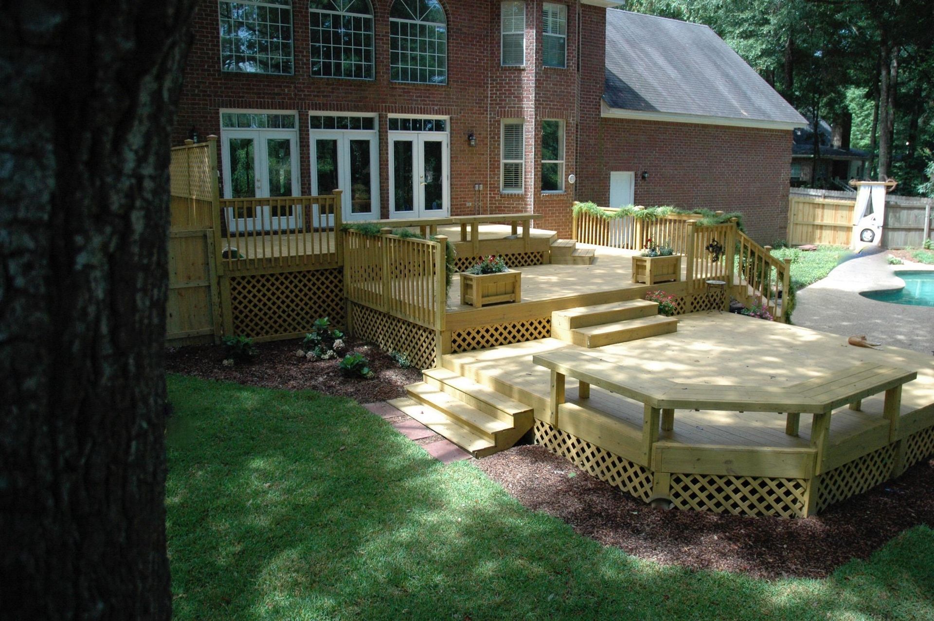 A multi-level wooden deck with lattice skirting and built-in benches, adjacent to a brick house and an outdoor pool.