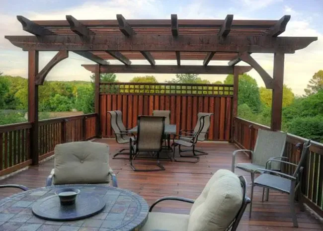 A dark wood pergola on a wooden deck featuring a dining table with chairs and a privacy fence, overlooking trees.