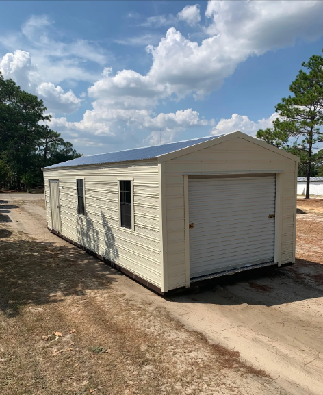 Tan metal garage with a roll-up door, side door, and two windows, on a gravel driveway under a partly cloudy sky. Tan metal garage with a roll-up door, side door, and two windows, on a gravel driveway under a partly cloudy sky.