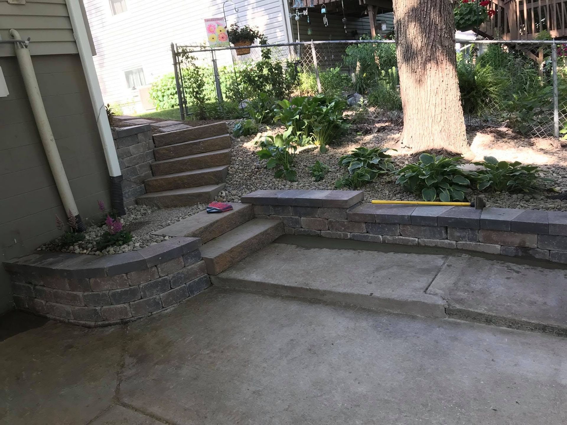 Concrete patio with steps, stone retaining wall, and a tree in a small backyard garden