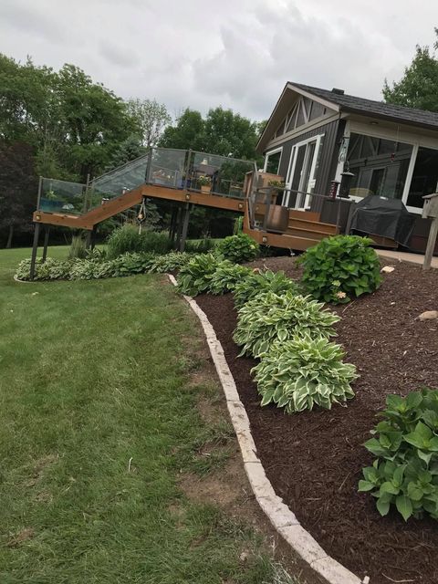 Landscaped yard with curved stone edging, mulch beds, shrubs, and a deck beside a house under cloudy skies