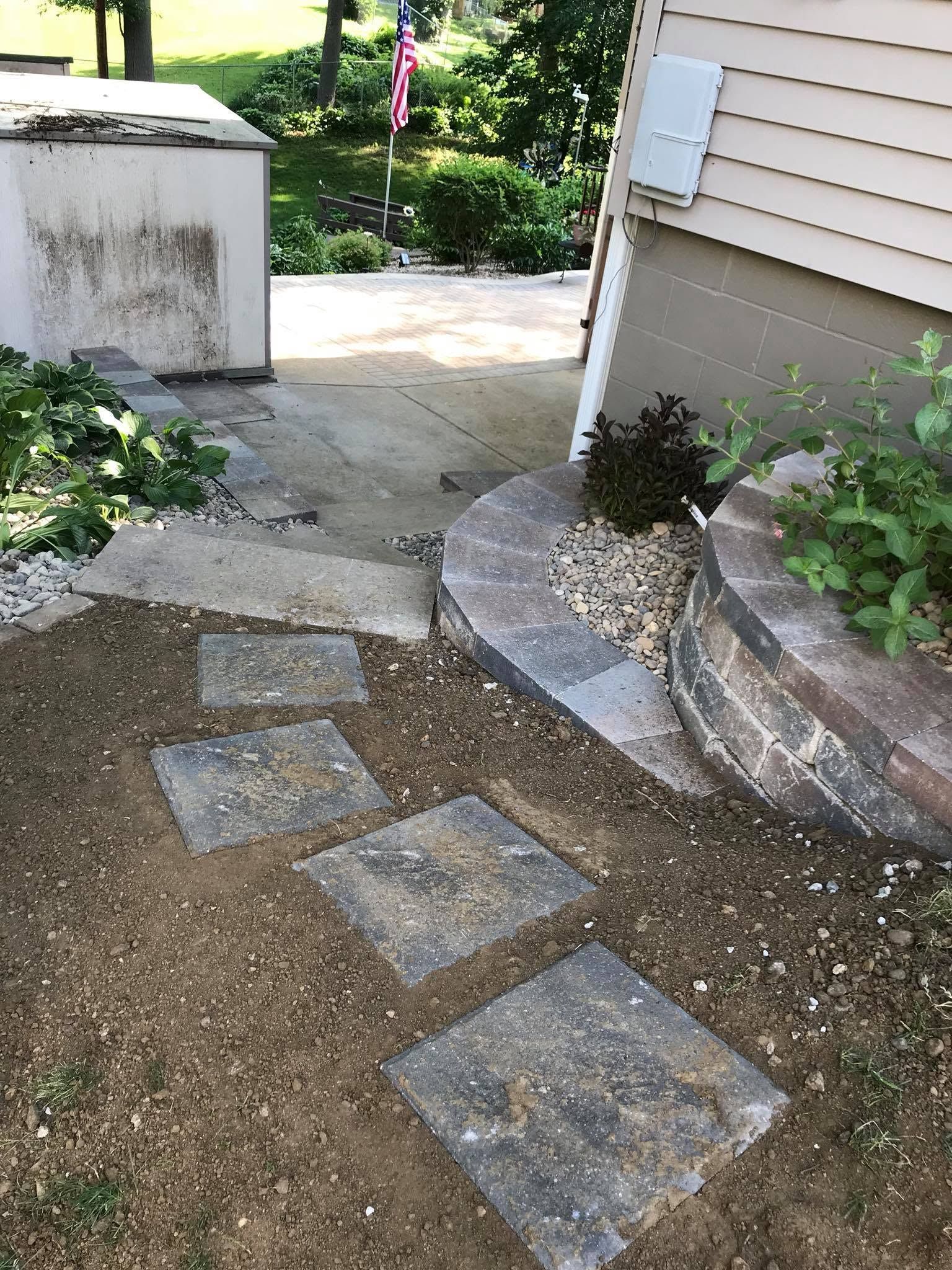 Stepping-stone path between a house wall and concrete patio, leading into a green backyard.