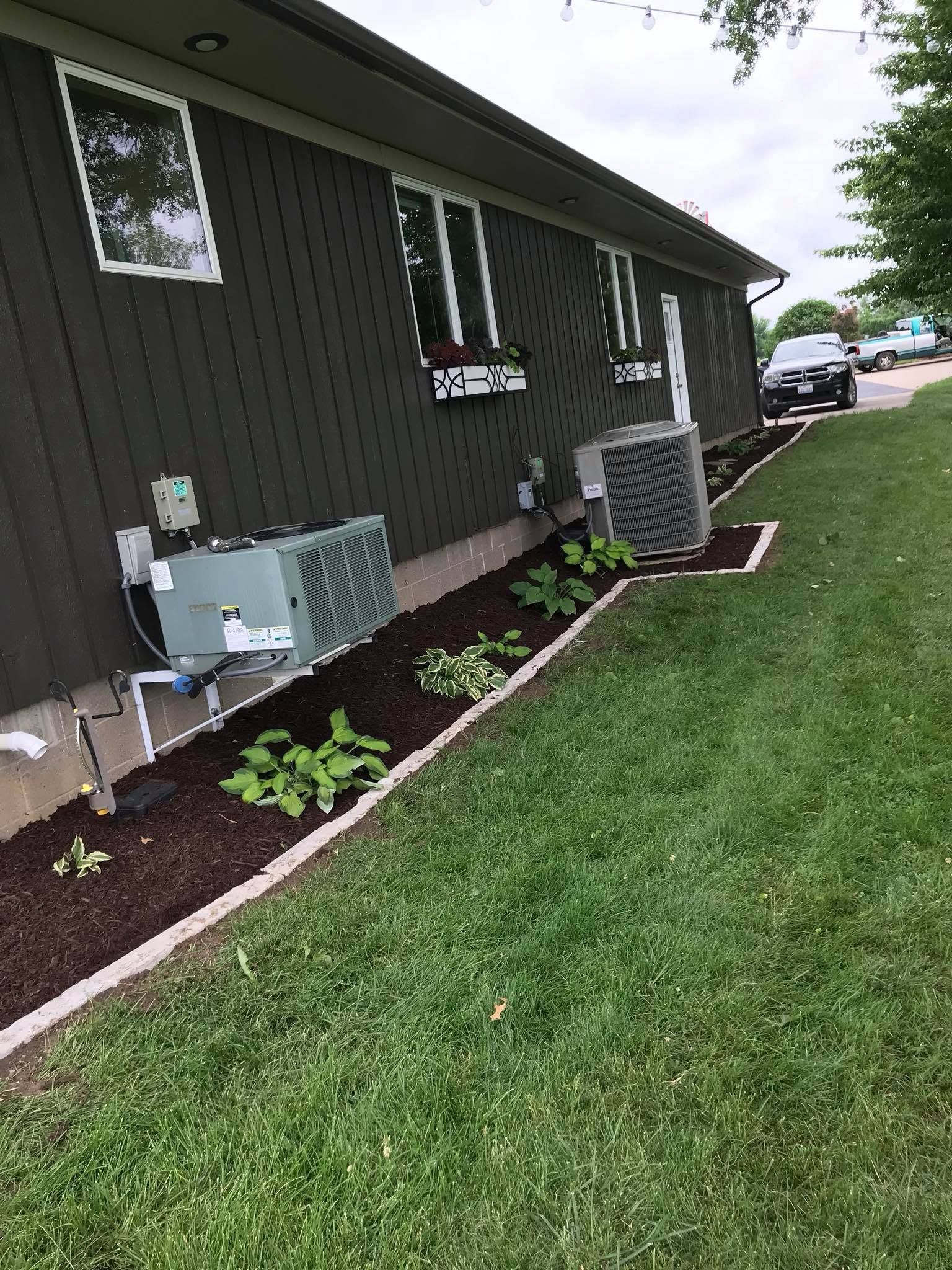 Side of a dark building with HVAC units, flower beds, and a grassy lawn beside a parked car row