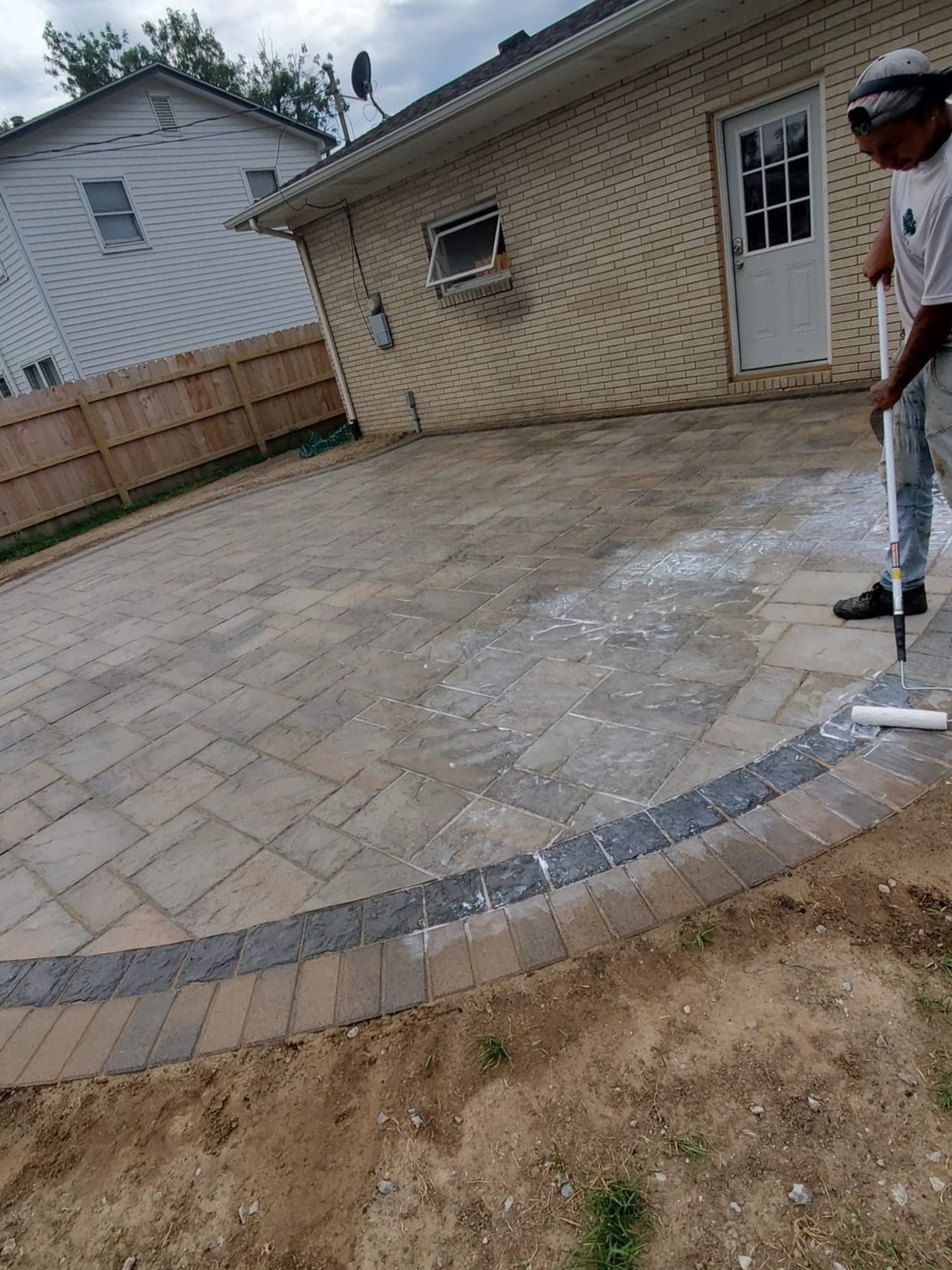 Person pressure-washing a curved stone patio beside a beige house under a cloudy sky