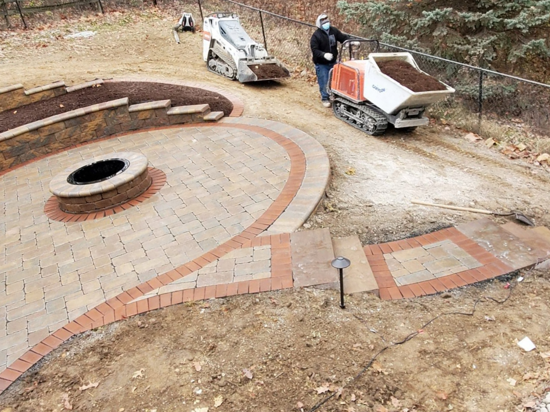 Curved stone walkway in a wooded area with a small circular opening and scattered construction materials