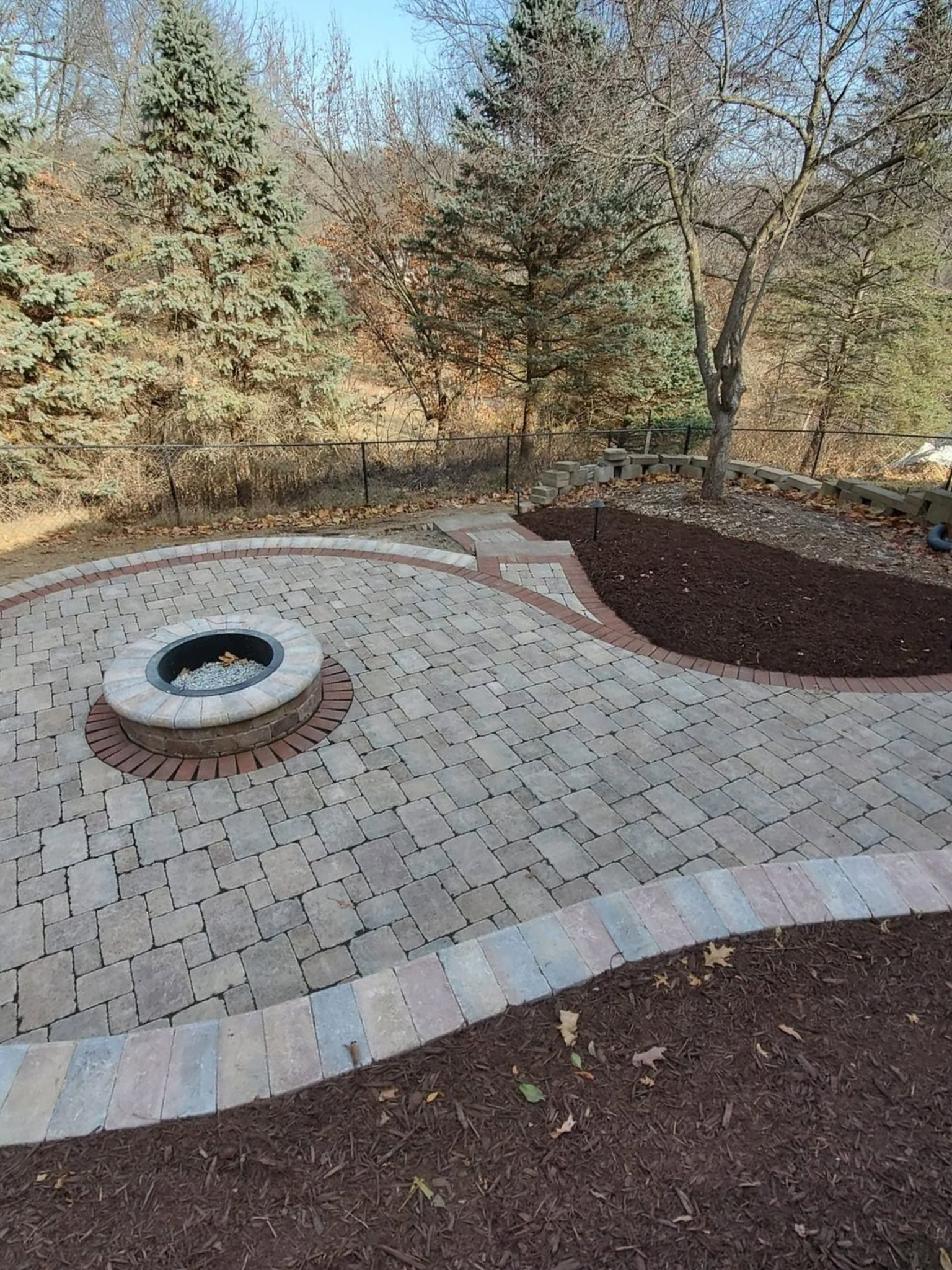 Curved brick-paved landscape feature with a circular drain, bordered by mulch and trees under a blue sky