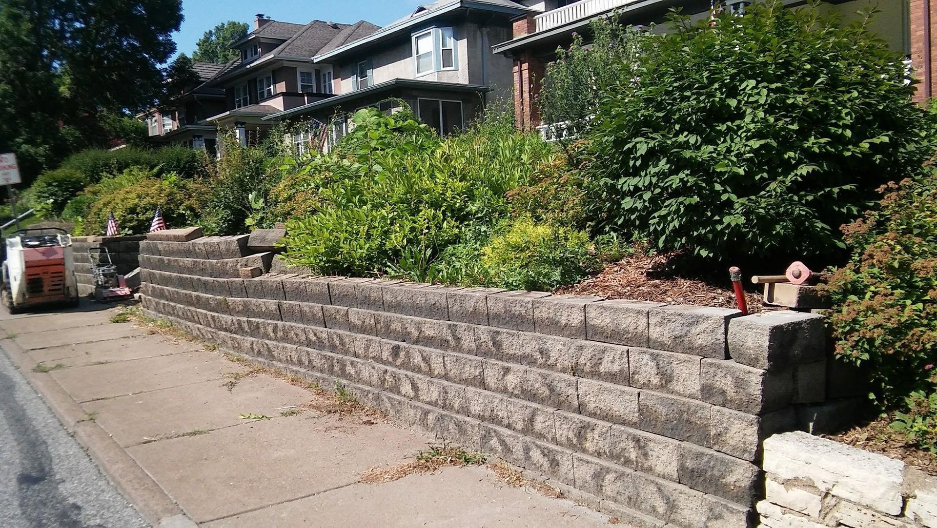 Stone retaining wall along a sidewalk beside a leafy garden and houses on a sunny street