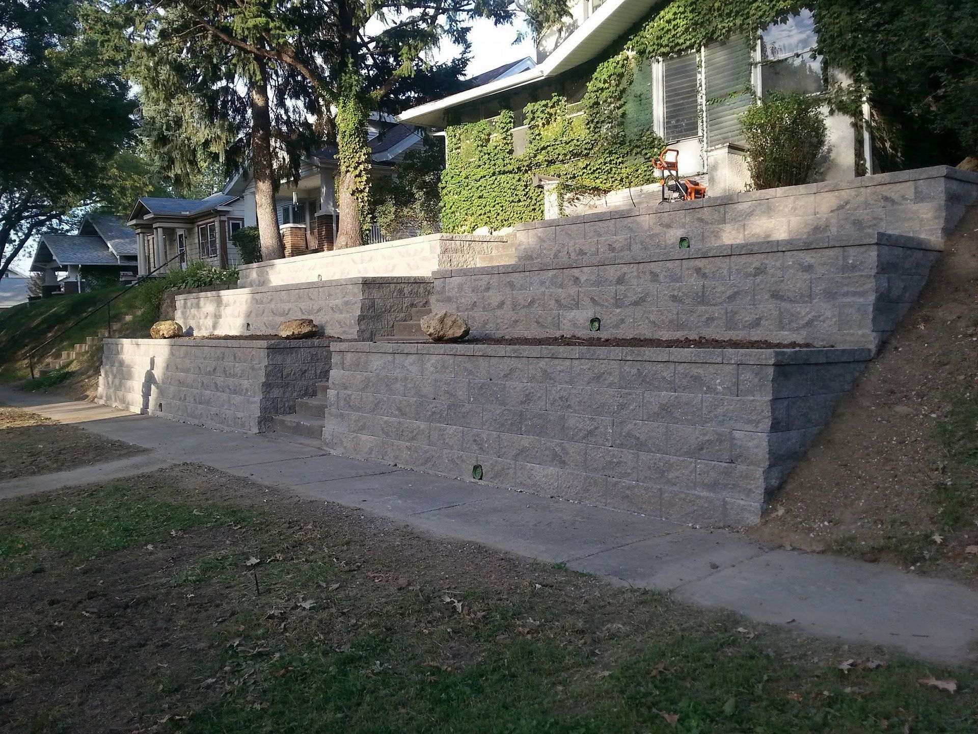 Tiered stone retaining wall with steps in a sloped yard beside a house.