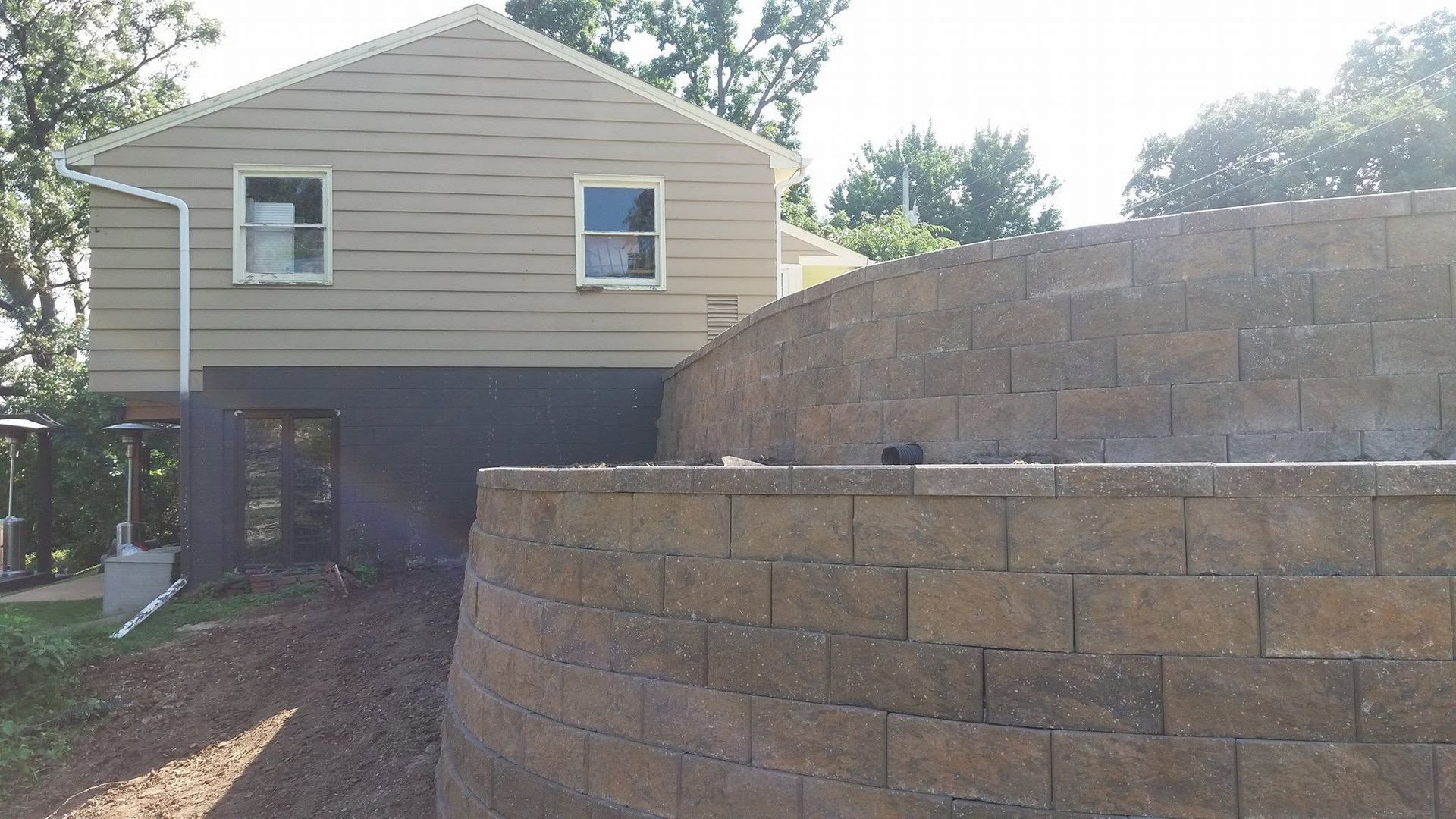 House with tan siding behind a large stacked-stone retaining wall and driveway