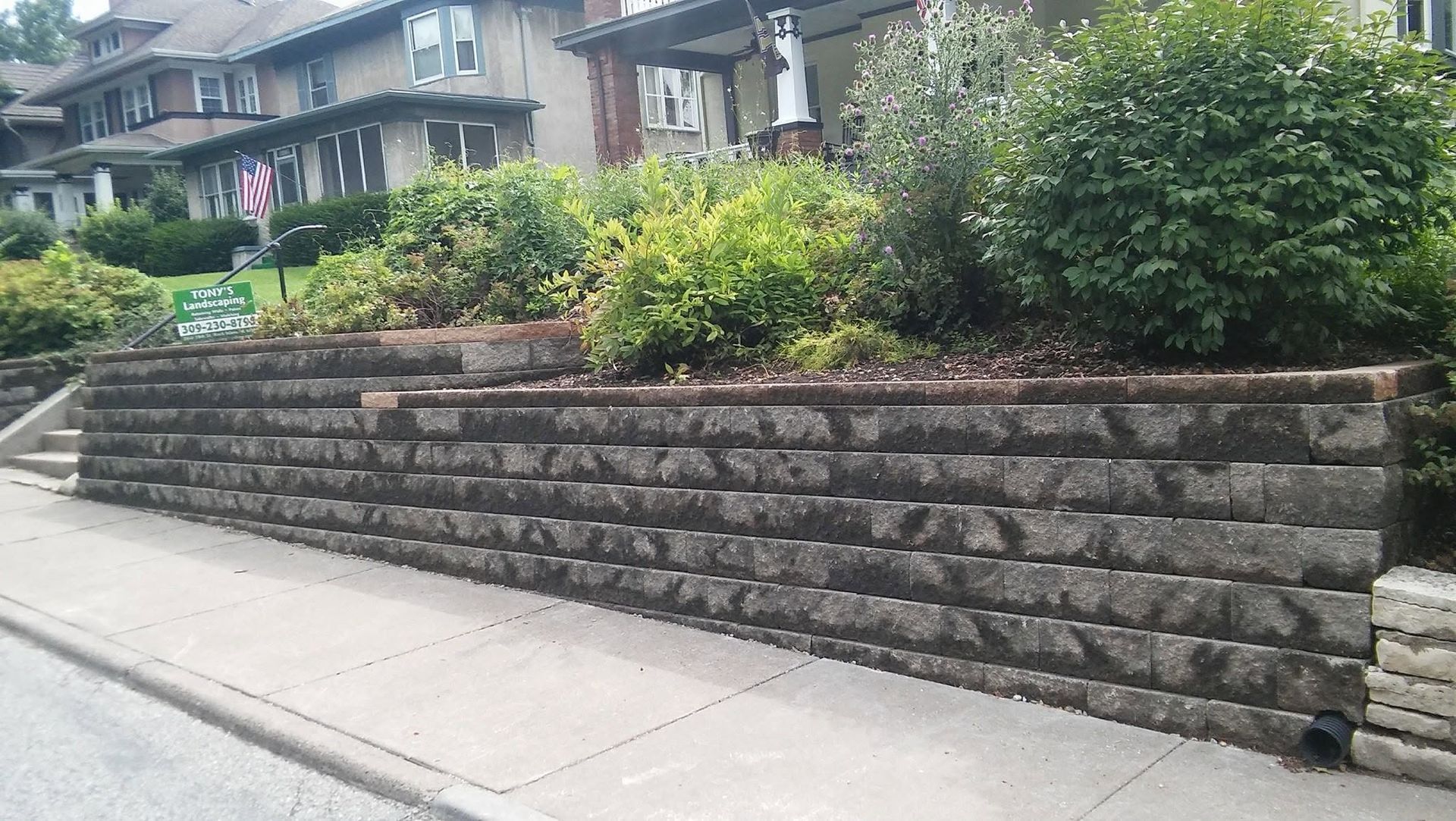 Stone retaining wall with shrubs along a residential sidewalk in front of houses