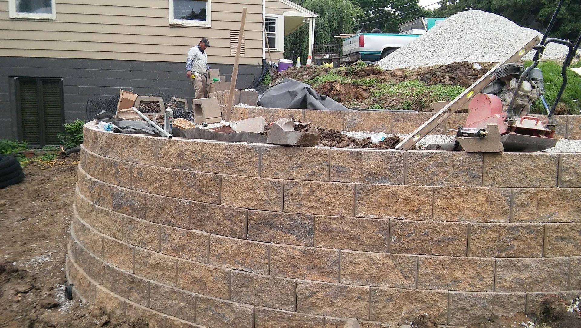 Workers building a curved stone retaining wall beside a house, with gravel piles and construction materials nearby