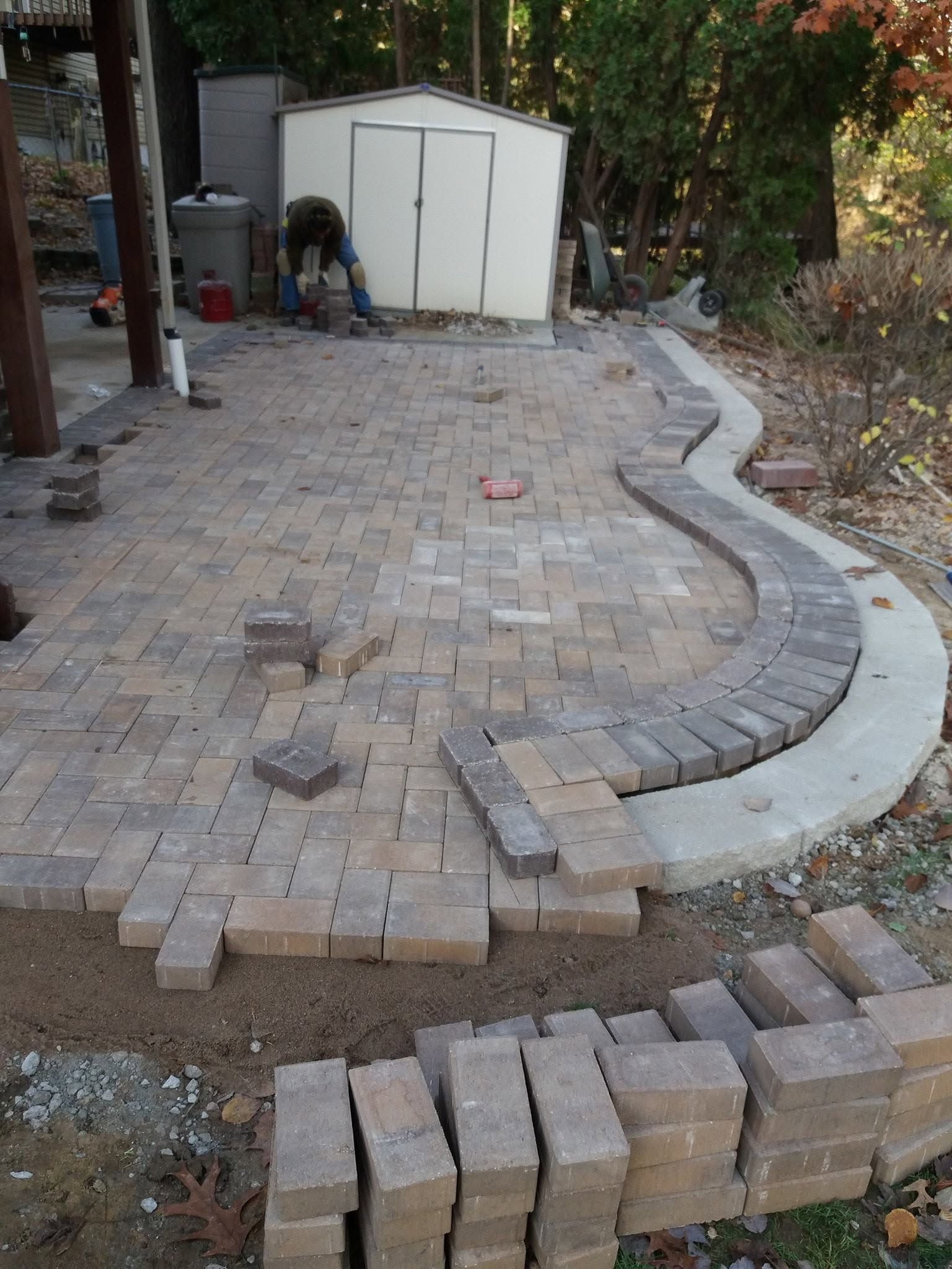 Partially paved brick patio under construction beside a shed, with stacked bricks and a curved concrete border.