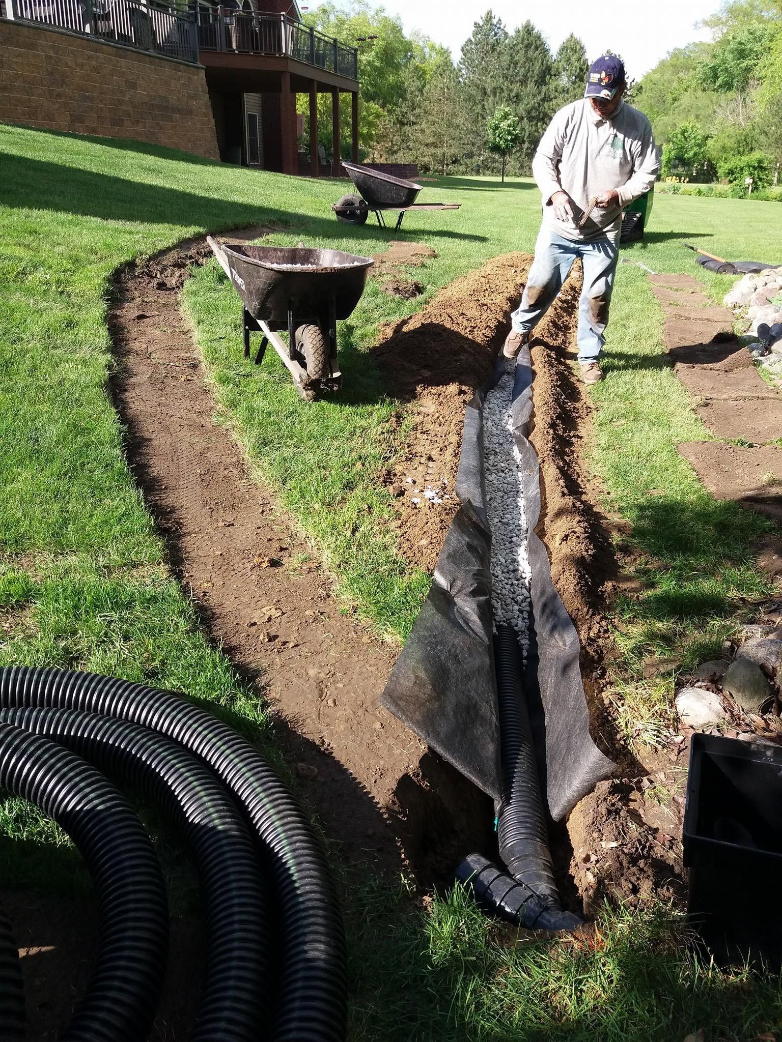 Worker standing beside a black plastic-lined trench in a grassy yard, with coiled pipe and a wheelbarrow nearby