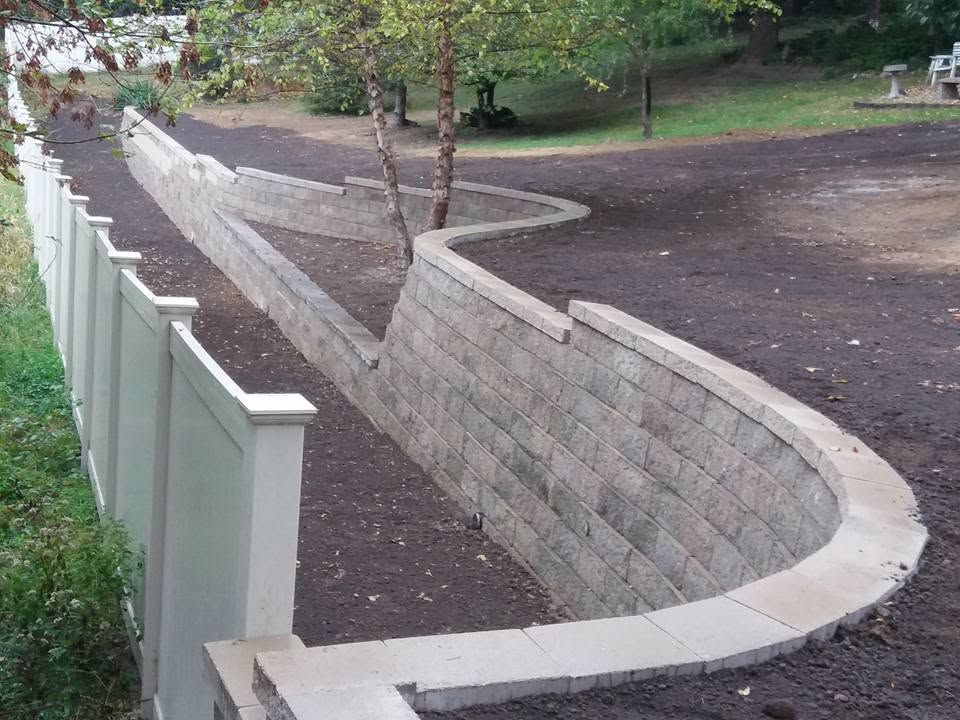 Curved concrete retaining wall beside a dirt path and white fence in a park-like area