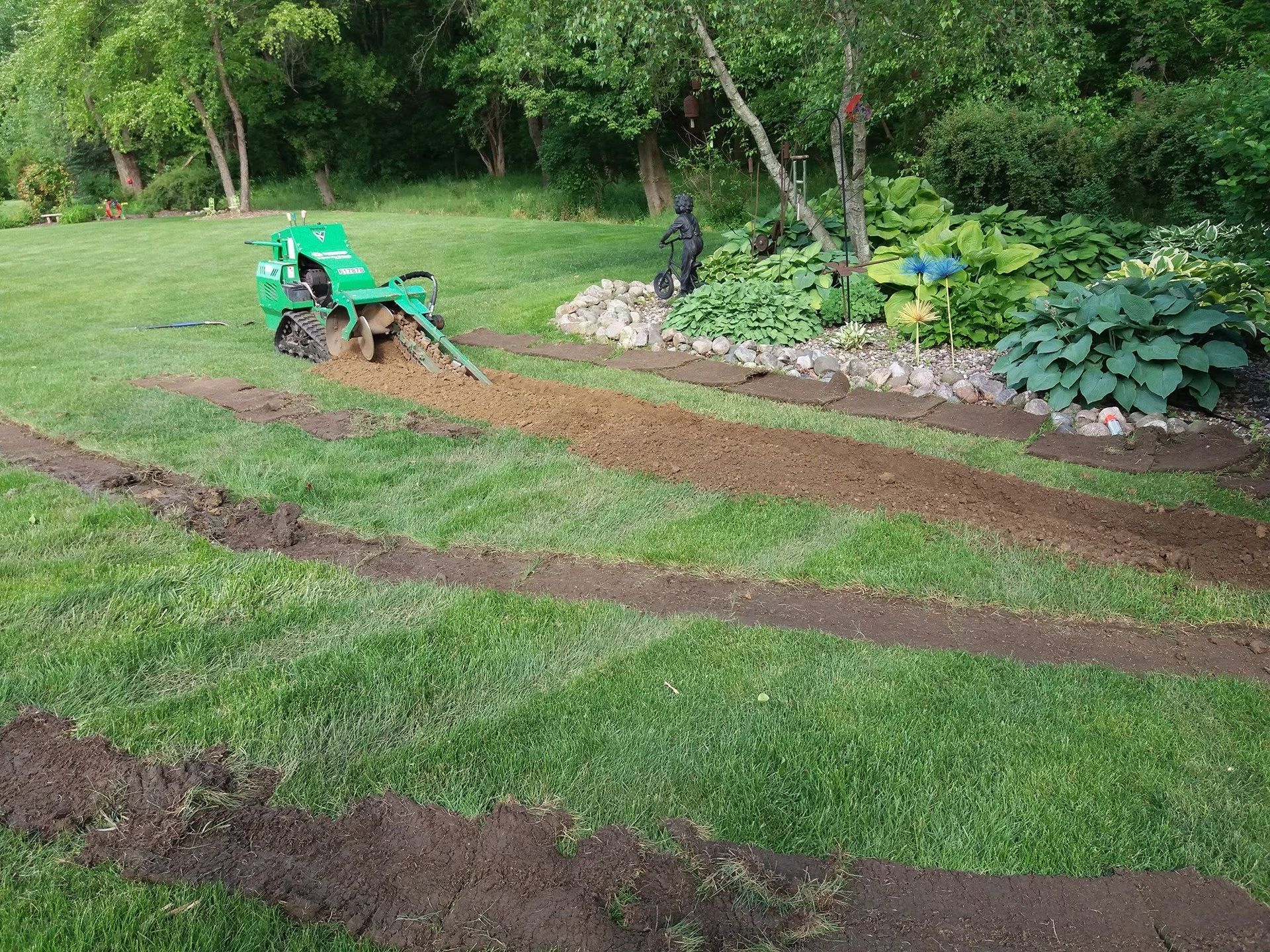 Green lawn with muddy tire tracks, a small tractor, and a person near a stone-lined garden bed.