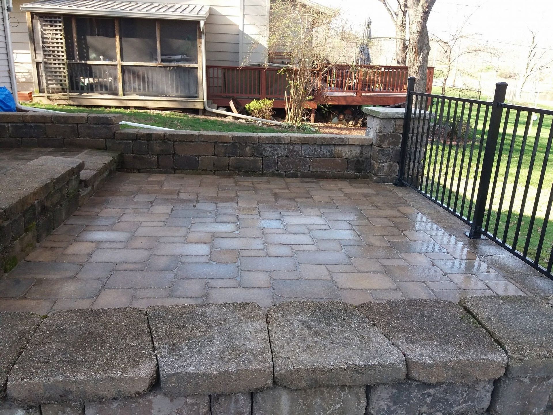 Stone-paver patio bordered by low retaining walls and black metal fence beside a house and deck.