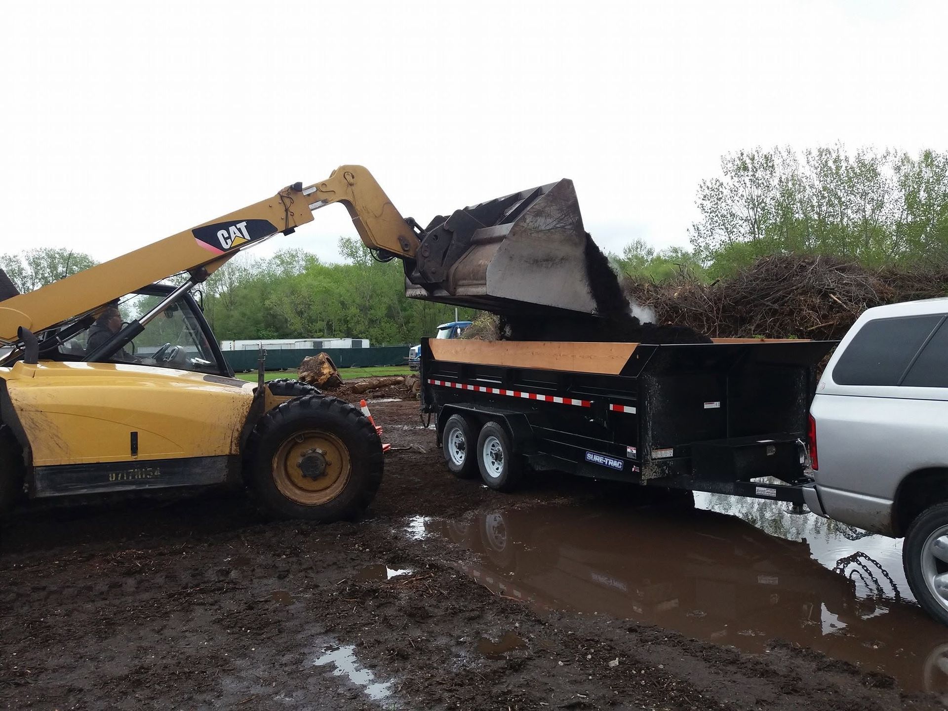 Yellow excavator loading debris into a black trailer beside a silver truck on a muddy roadside