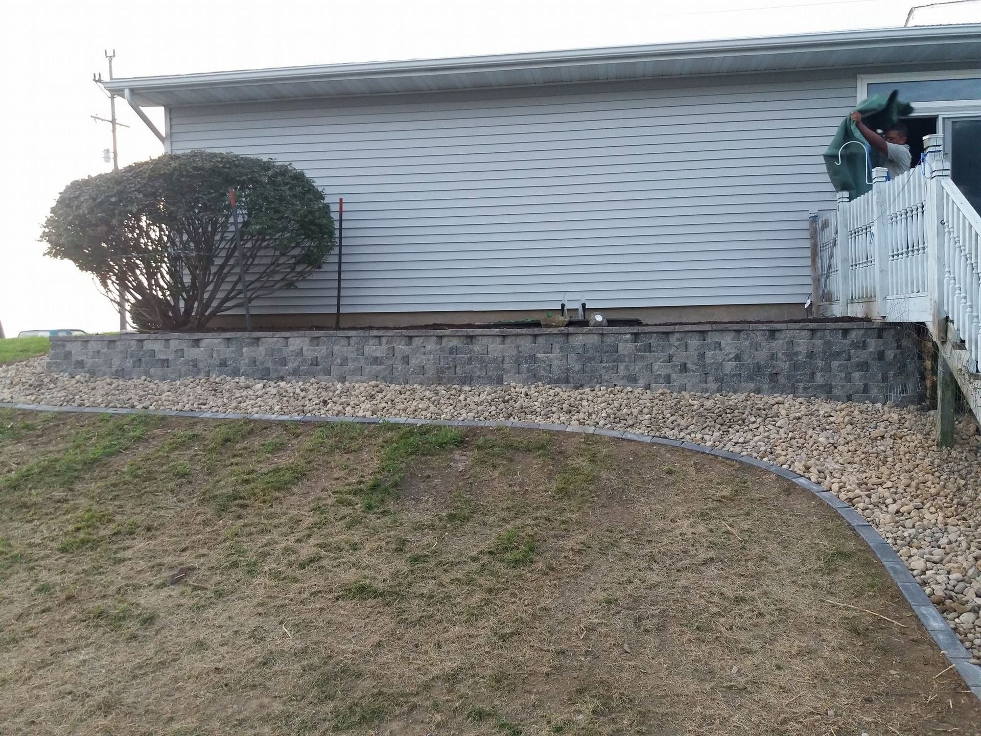 House with sloped yard, stone retaining wall, shrubs, and a person on the porch stairs