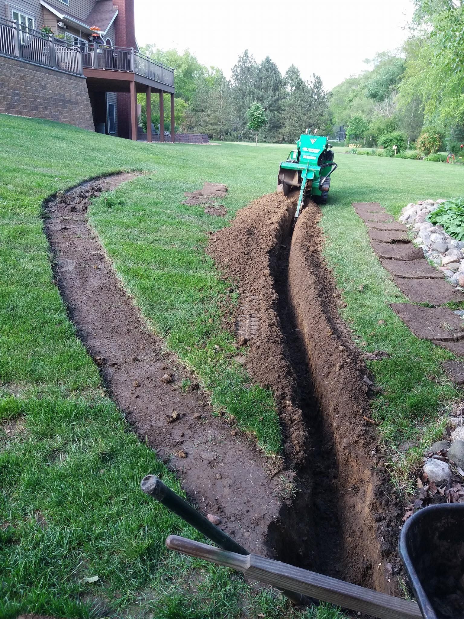 Green machine digging a muddy trench through a grassy yard near a house