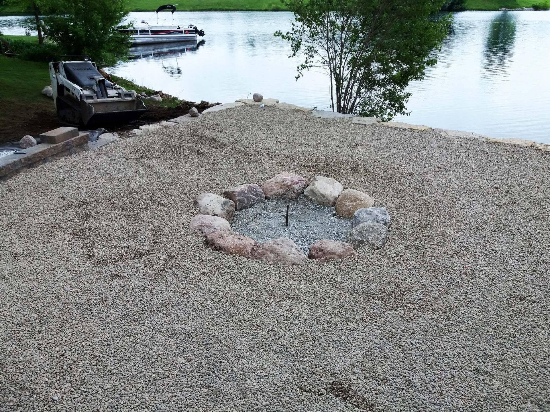 Gravel fire pit beside a calm lakeside with a dock and boat in the background