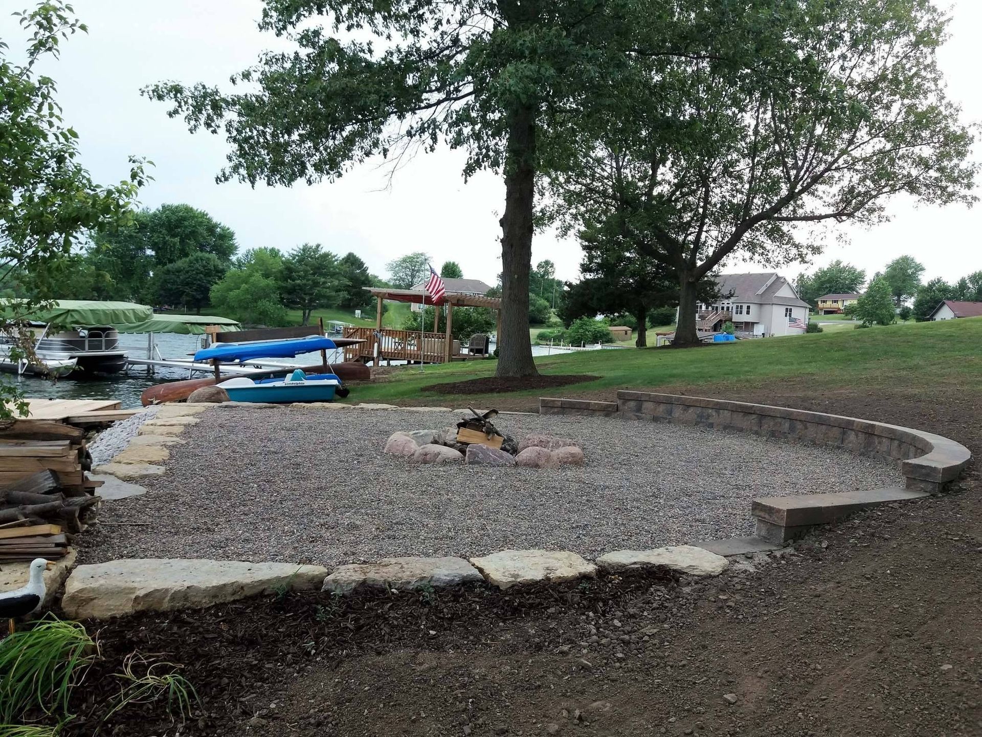 Rock-lined lakeside gravel area with benches, trees, and a blue boat dock nearby