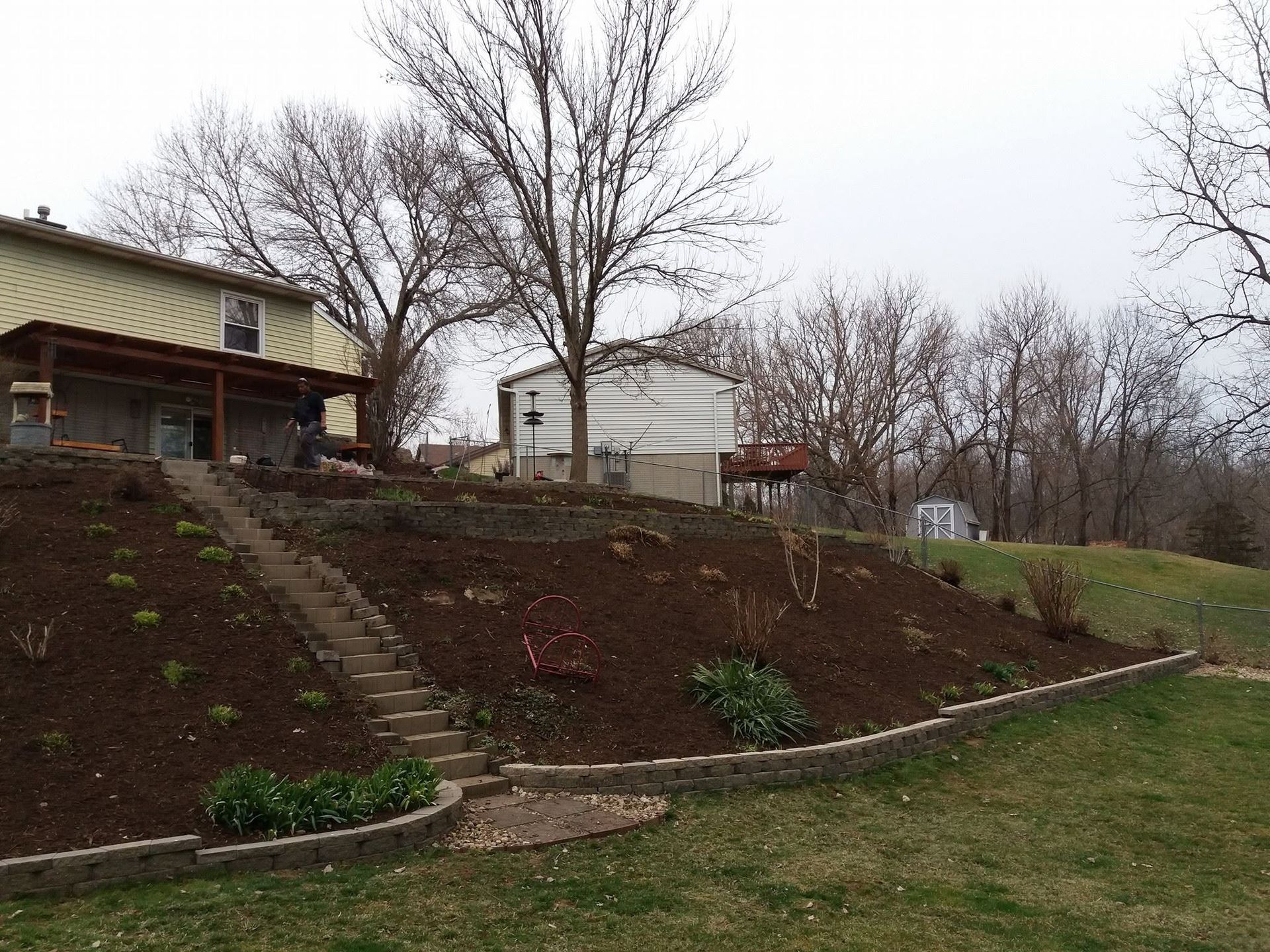 Rural hillside home with steps, bare trees, and a grassy yard on an overcast day