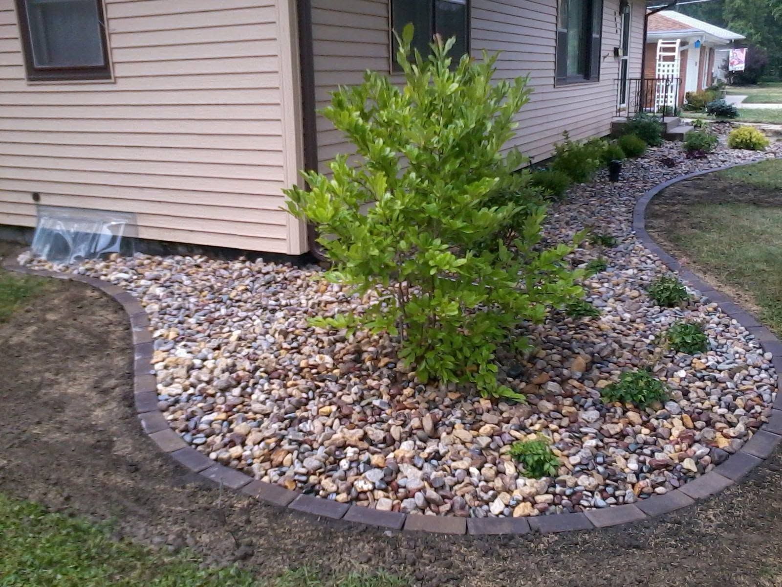 Curved stone landscape border around a green shrub beside a beige house and walkway