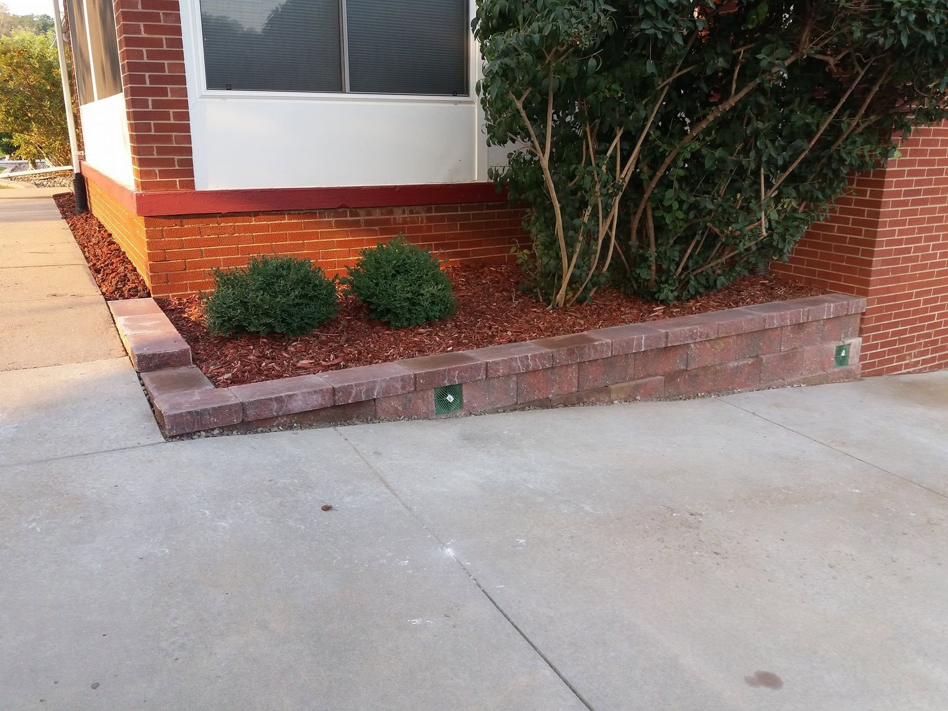 Brick house corner with a concrete driveway and small landscaped shrubs in front