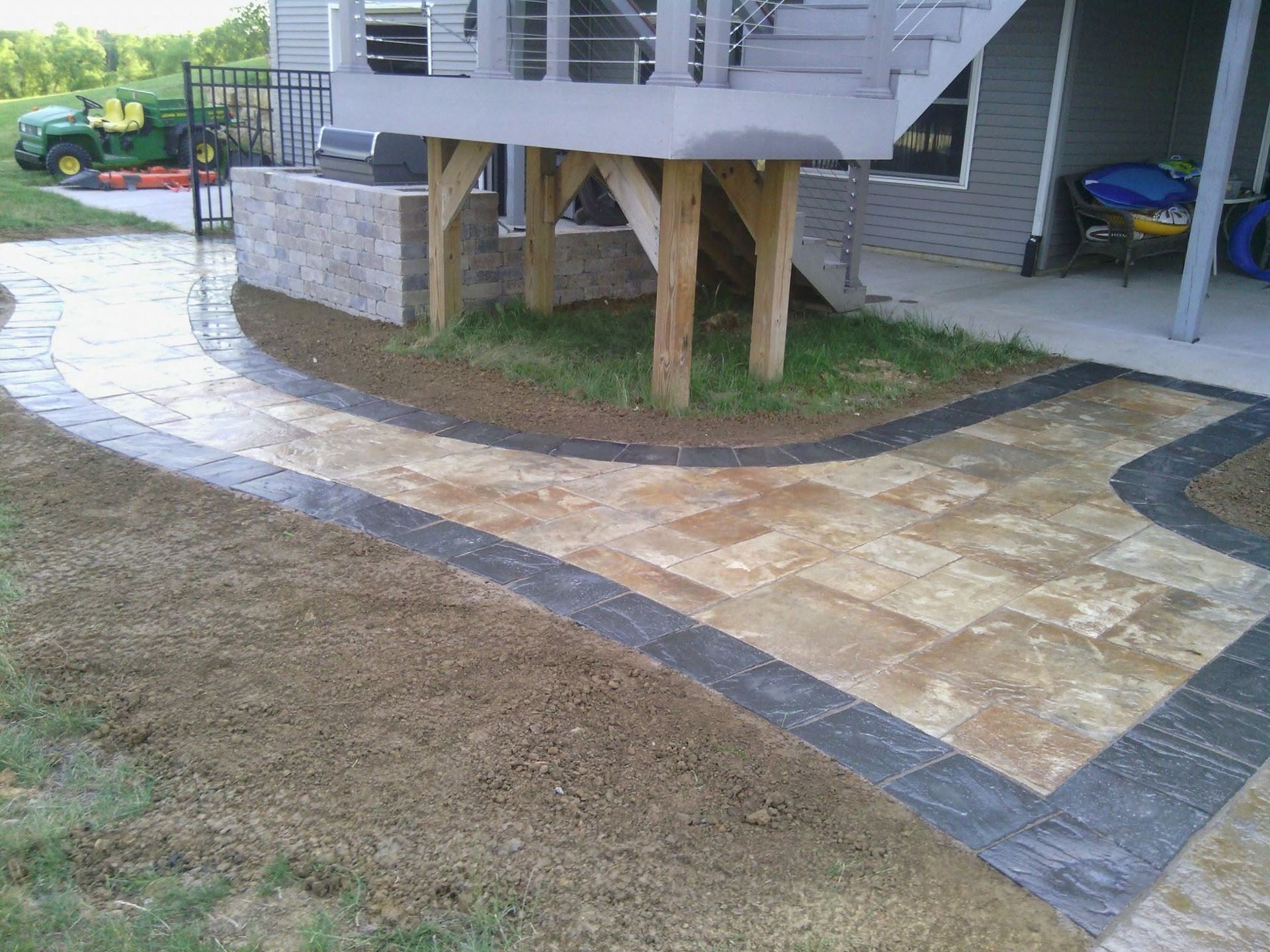 Stone walkway curves around a raised wooden deck beside a house and driveway.